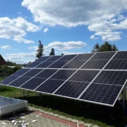 Panel słoneczny ustawiony na trawie obok domu z czerwonym dachem i błękitnym niebem w tle, dodatkowe panele na palecie leżą na ziemi.