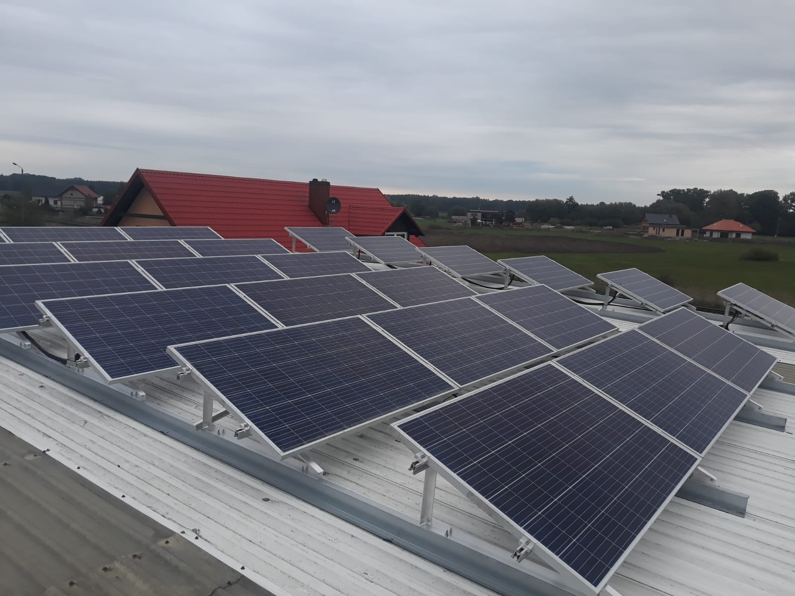 Rząd paneli słonecznych na białym dachu z widokiem na czerwony dach domu i zielone pola w tle, pochmurne niebo.
