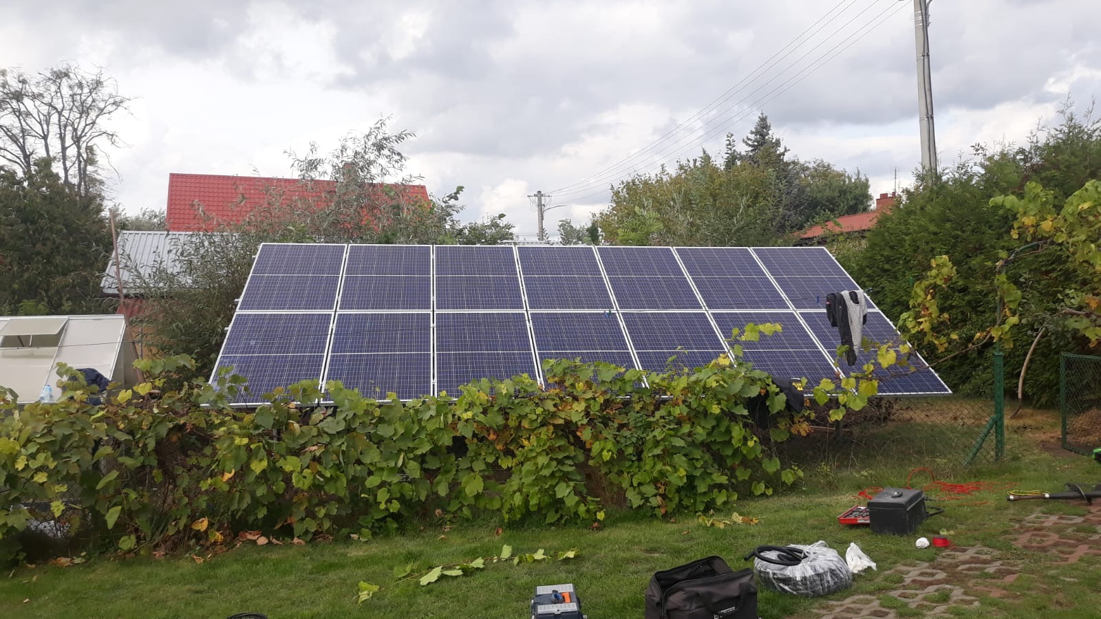 Panele słoneczne na trawniku przed domem, częściowo zasłonięte krzewami winorośli, widoczne narzędzia i torba monterska.