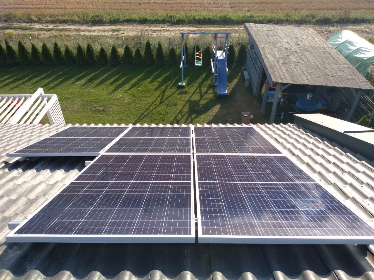 Panele słoneczne zamontowane na szarym, falistym dachu domu jednorodzinnego, w tle zielony trawnik, rząd tuj i pole uprawne.