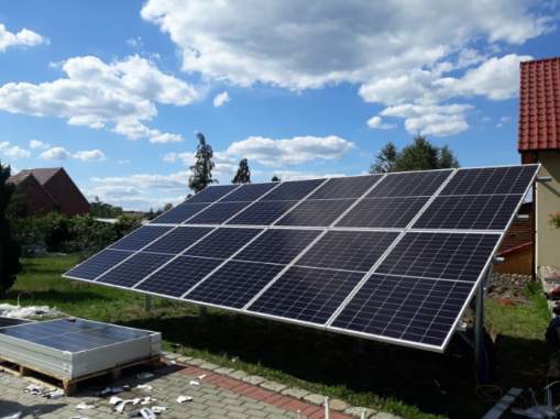 Panel słoneczny ustawiony na trawie obok domu z czerwonym dachem i błękitnym niebem w tle, dodatkowe panele na palecie leżą na ziemi.
