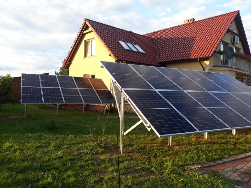 Panele fotowoltaiczne na trawniku przed domem jednorodzinnym z czerwoną dachówką, widoczne w popołudniowym słońcu.