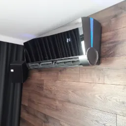 Nowoczesny klimatyzator z podświetleniem LED na drewnianej ścianie, wyświetlający temperaturę 21 stopni, obok czarna zasłona.