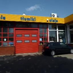 Budynek warsztatu samochodowego z czerwonymi drzwiami garażowymi, żółtym dachem z niebieskimi napisami 'Tłumiki' i 'Oleje', oraz zaparkowanym czarnym samochodem osobowym.