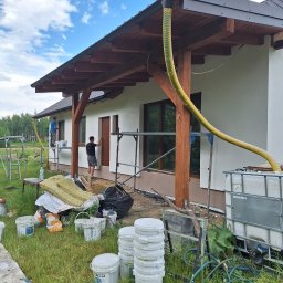 Bart. Bud - Dwóch pracowników przy pracy nad elewacją domu jednorodzinnego: jeden maluje ramę okienną, drugi pracuje na rusztowaniu, z żółtym wężem doprowadzającym materiał do urządzenia do natrysku, widoczne...