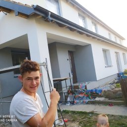 Bart. Bud - Młody mężczyzna z uśmiechem na twarzy, ubrudzony farbą, stoi na tle domu w trakcie malowania elewacji, widoczne rusztowanie i zabezpieczone okna.