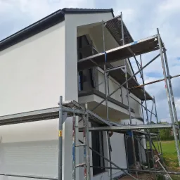 Nowoczesny dom z białą elewacją, widok z dołu na rusztowanie. Świeżo otynkowana ściana i widoczne elementy wykończeniowe.