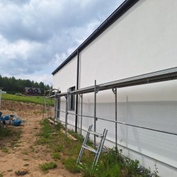 Bart. Bud - Świeżo pomalowana, biała elewacja budynku z czarnymi rynnami, widoczna ze rusztowania w Mrągowie. W tle zielony krajobraz i pochmurne niebo.