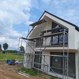 Bart. Bud - Nowoczesny dom z białą elewacją w trakcie prac. Widoczne rusztowania, okna zabezpieczone folią. W tle zielone pola i błękitne niebo z chmurami.