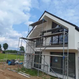 Nowoczesny dom z białą elewacją w trakcie prac. Widoczne rusztowania, okna zabezpieczone folią. W tle zielone pola i błękitne niebo z chmurami.