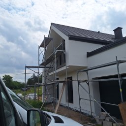 Bart. Bud - Nowoczesny dom jednorodzinny z białą elewacją w trakcie prac wykończeniowych. Widoczne rusztowania i fragment samochodu dostawczego. Tło: zielone drzewa i pochmurne niebo.