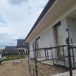 Bart. Bud - Fragment elewacji domu w trakcie tynkowania, okna zabezpieczone folią, widoczne rusztowania i domy w tle. Ujęcie z dołu, pochmurne niebo.