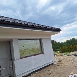 Bart. Bud - Dom jednorodzinny w trakcie tynkowania elewacji. Okno zabezpieczone folią. Widoczne rusztowanie i teren budowy. Dach pokryty czarną dachówką.