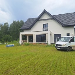 Bart. Bud - Nowoczesny dom jednorodzinny z białą elewacją i ciemną dachówką. Na pierwszym planie trawnik i samochód dostawczy z otwartymi drzwiami.