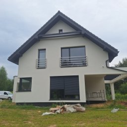 Bart. Bud - Nowoczesny dom jednorodzinny z jasną elewacją, czarnym dachem i ramami okiennymi. Widoczne rusztowanie i materiały budowlane sugerują prace wykończeniowe.