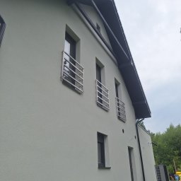 Bart. Bud - Jasna elewacja domu z trzema oknami zabezpieczonymi metalowymi balustradami. Widok z dołu, fragment dachu i drzewa w tle.