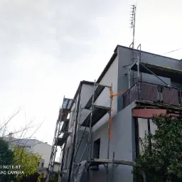 Dom jednorodzinny w trakcie prac elewacyjnych, widoczne rusztowania, świeżo pomalowana szara elewacja i antena na dachu.