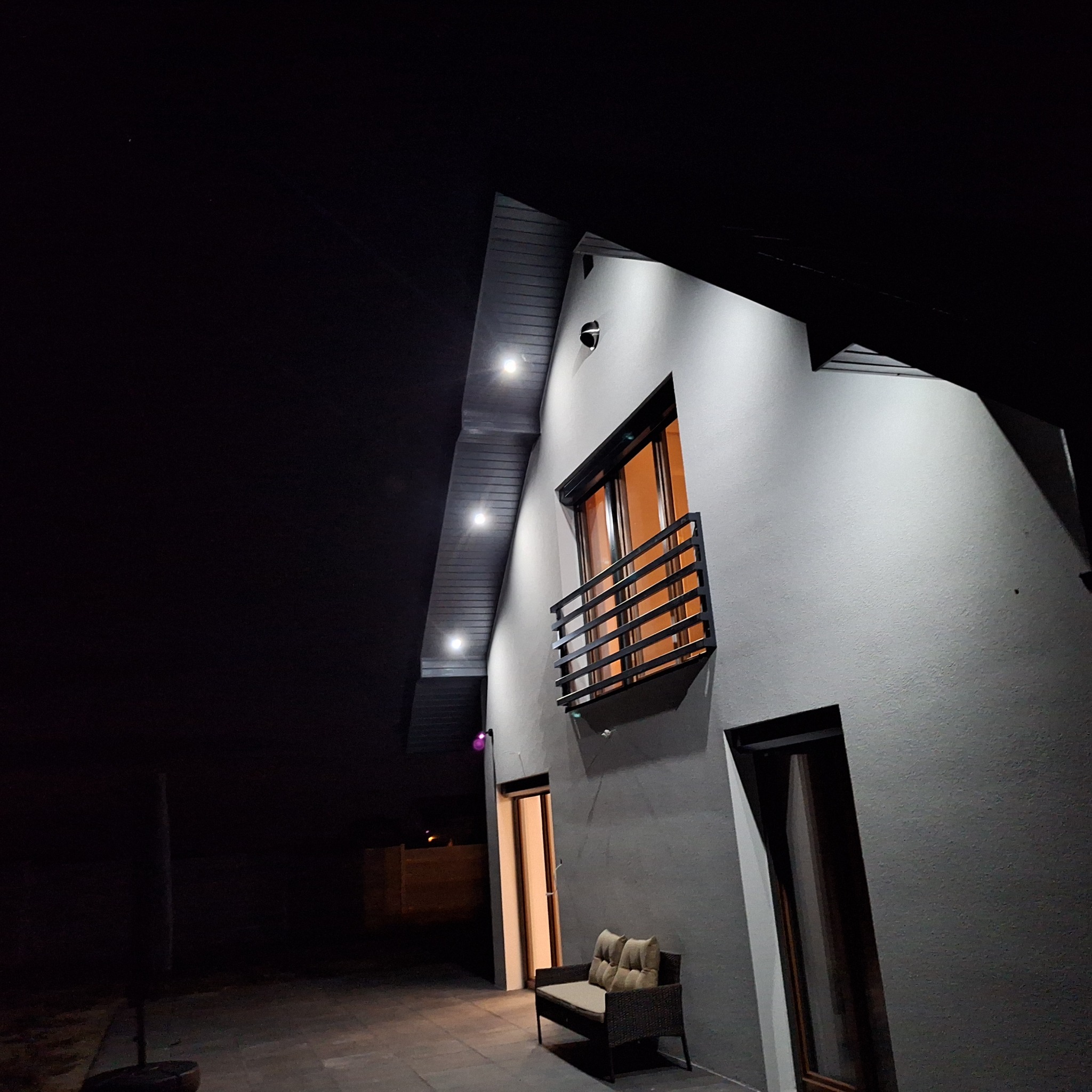 Nowoczesny dom z szarą elewacją oświetloną punktowymi lampami, widoczny balkon z czarną balustradą i rattanowa ławka na tarasie w nocnej scenerii.