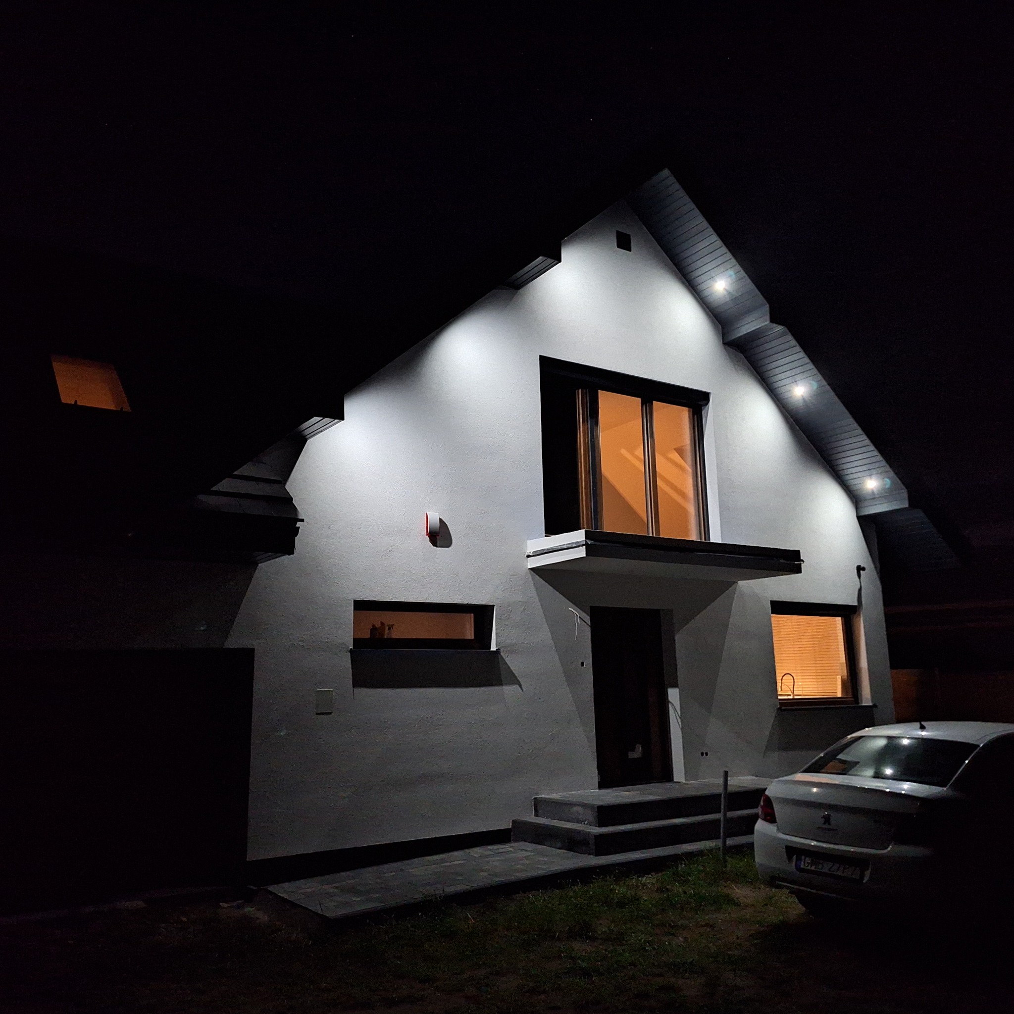 Nowoczesny dom jednorodzinny oświetlony nocą, z widoczną fakturą tynku i subtelnymi elementami dekoracyjnymi elewacji oraz zaparkowanym samochodem osobowym.