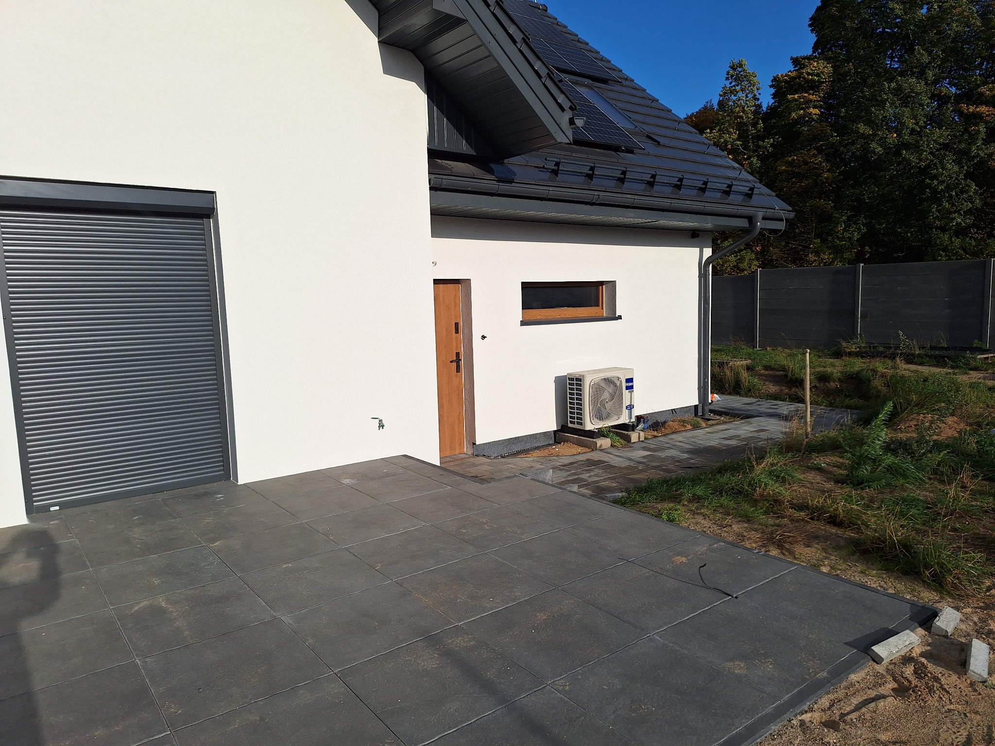 Nowoczesny dom z białą elewacją, grafitową stolarką okienną i roletami, panele słoneczne na dachu, klimatyzator na zewnątrz, wyłożony płytami taras.