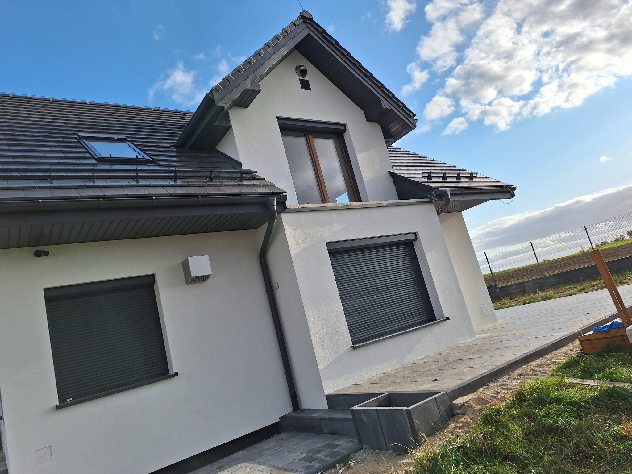 Nowoczesny dom jednorodzinny z białą elewacją, szarą dachówką i roletami zewnętrznymi, widok z boku na tle błękitnego nieba z chmurami.