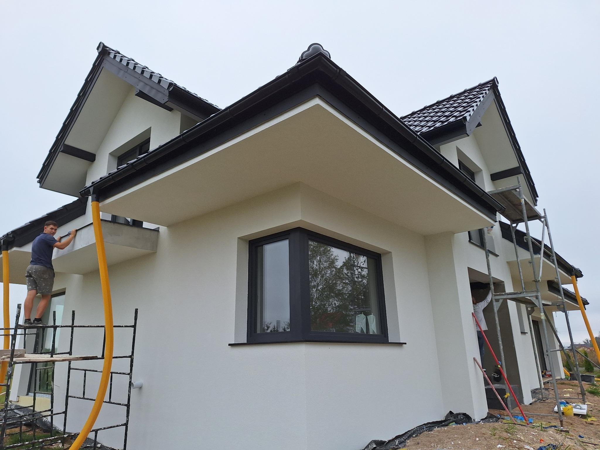 Pracownik na rusztowaniu wykańcza elewację nowoczesnego domu z antracytową stolarką okienną i dachem, widoczny żółty wąż do agregatu.
