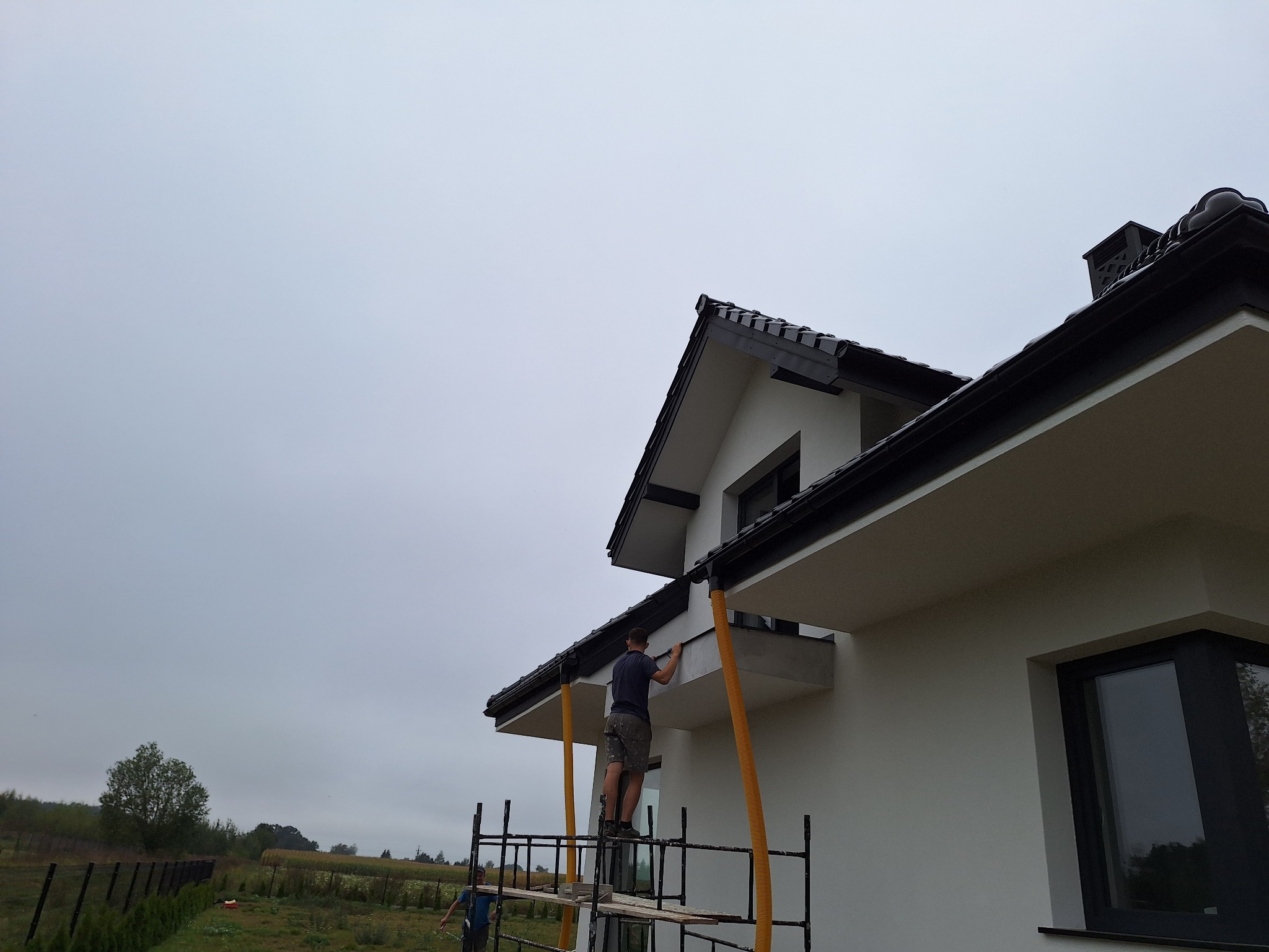 Pracownik na rusztowaniu w Mrągowie, poprawiający detale elewacji nowoczesnego domu z czarną stolarką okienną i dachem, widok z dołu na tle pochmurnego nieba.