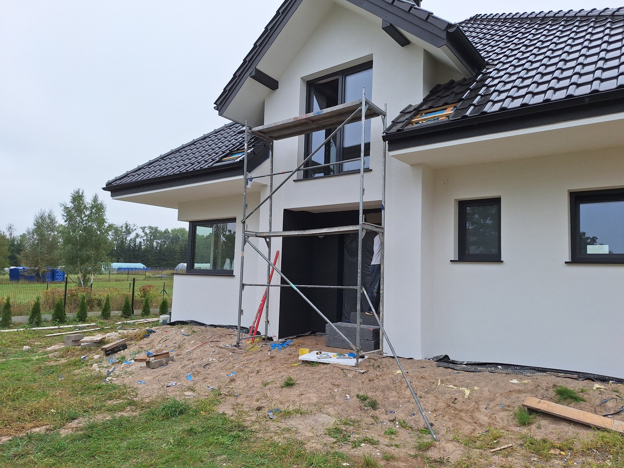 Nowoczesny dom jednorodzinny z białą elewacją, czarną stolarką okienną i ciemną dachówką, w trakcie prac elewacyjnych z użyciem rusztowania.