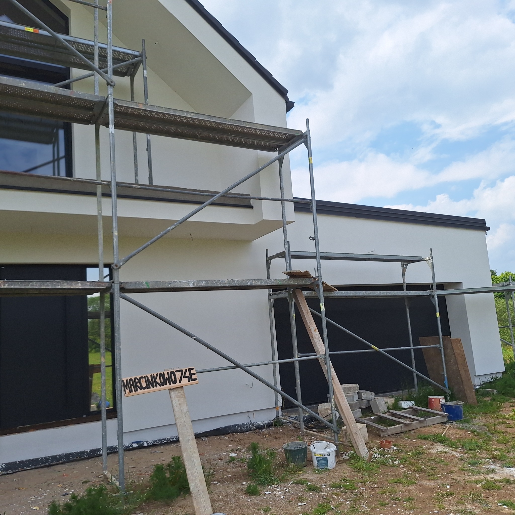 Biała elewacja domu jednorodzinnego w trakcie prac wykończeniowych, widoczne rusztowania, tabliczka z adresem i materiały budowlane.