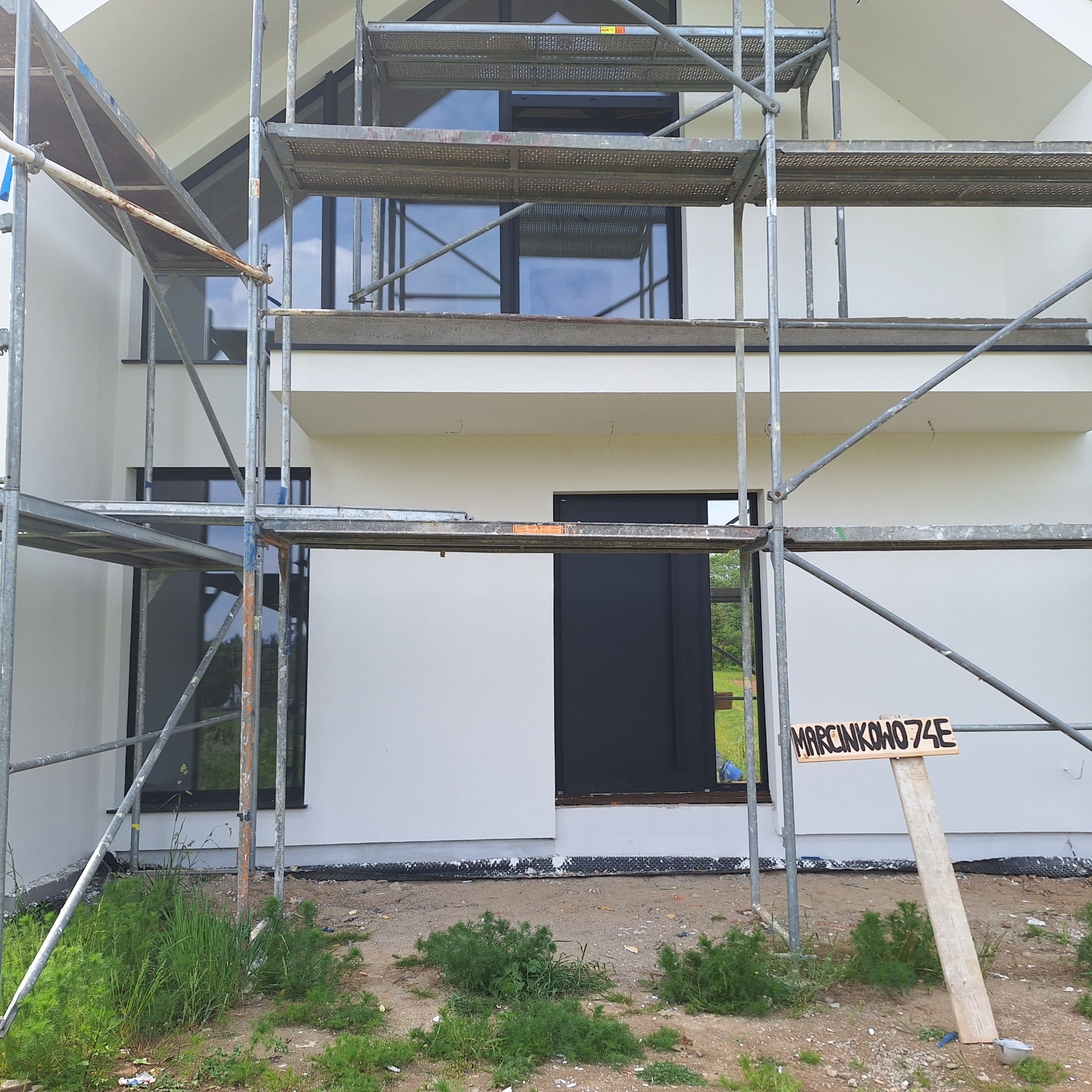 Świeżo pomalowana, biała elewacja domu jednorodzinnego z czarnymi oknami, widoczna spod rusztowania. Na dole tabliczka z adresem.