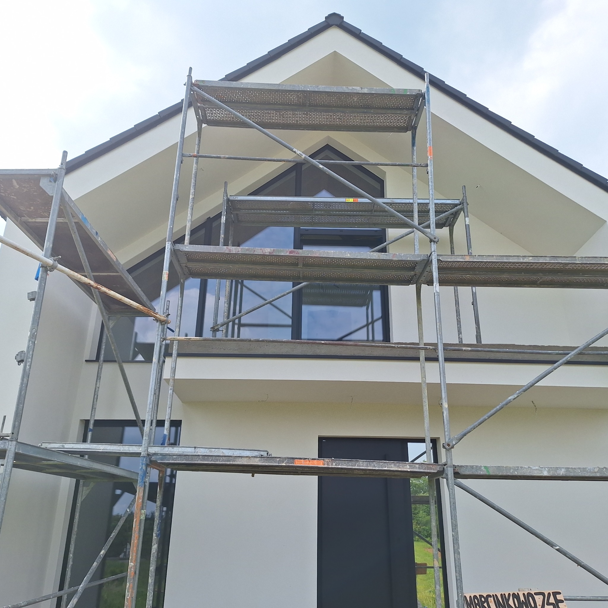 Nowoczesny dom jednorodzinny w trakcie prac elewacyjnych. Widoczne rusztowania i świeżo pomalowana, jasna elewacja z czarnymi akcentami okien i drzwi.