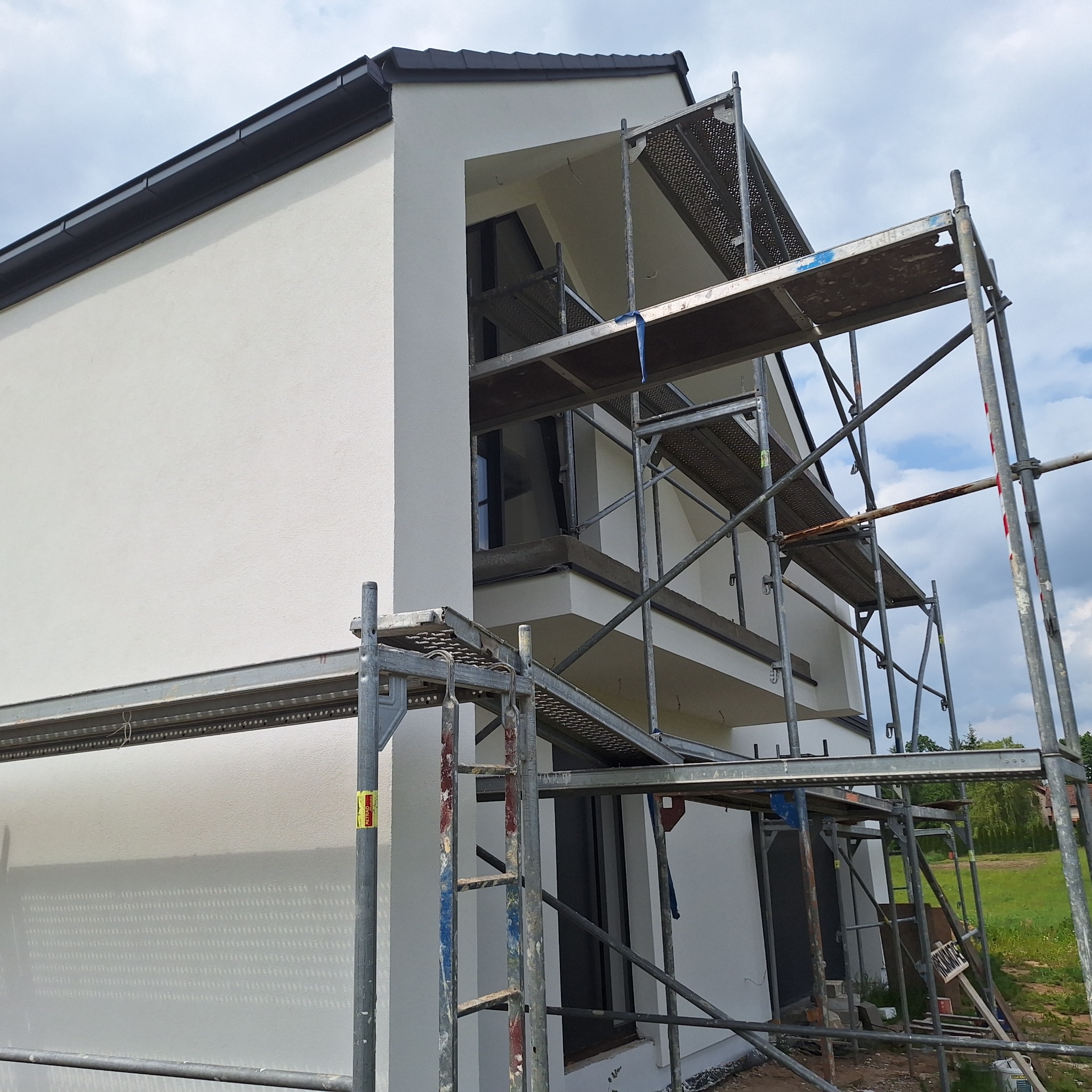 Nowoczesny dom z białą elewacją, widok z dołu na rusztowanie. Świeżo otynkowana ściana i widoczne elementy wykończeniowe.