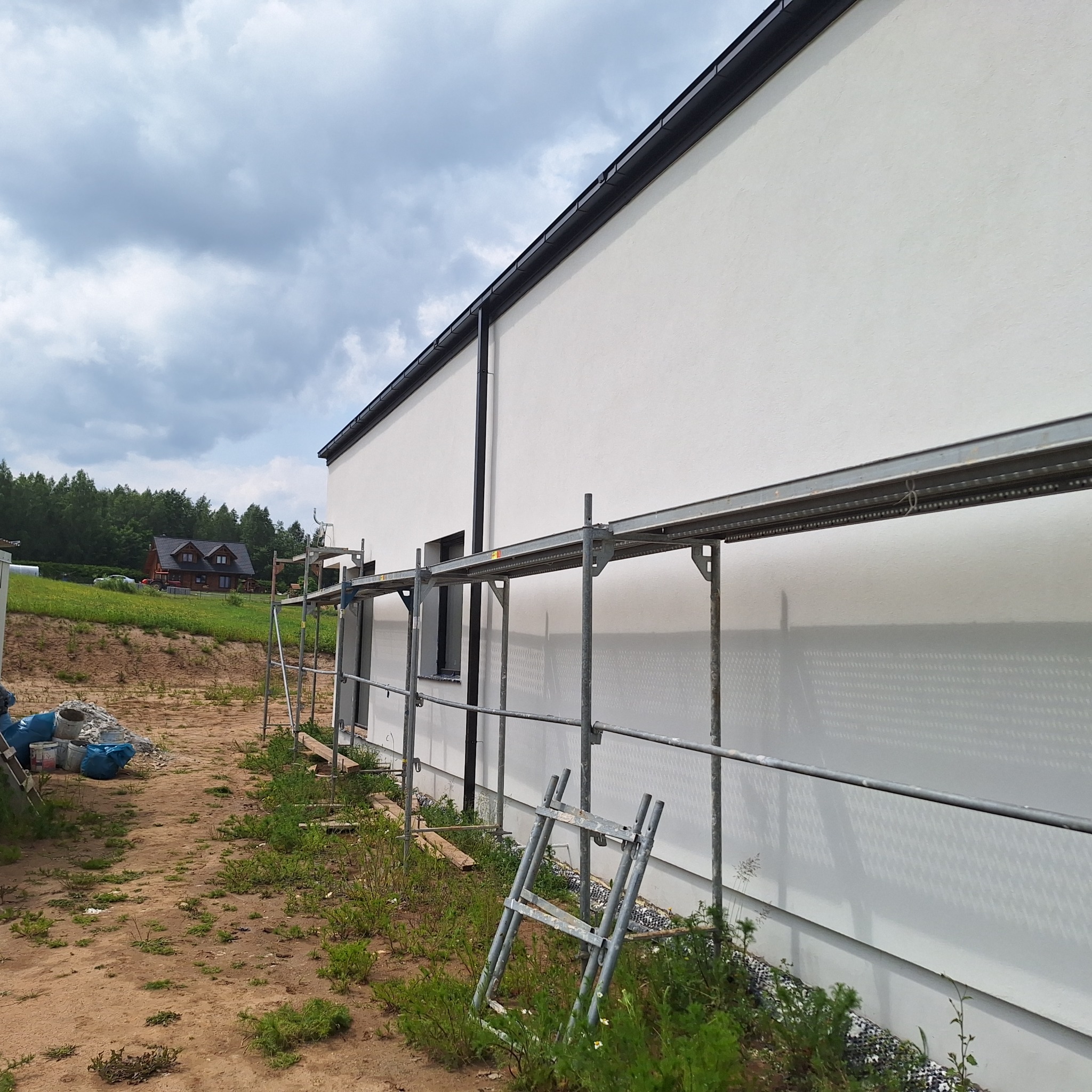 Świeżo pomalowana, biała elewacja budynku z czarnymi rynnami, widoczna ze rusztowania w Mrągowie. W tle zielony krajobraz i pochmurne niebo.