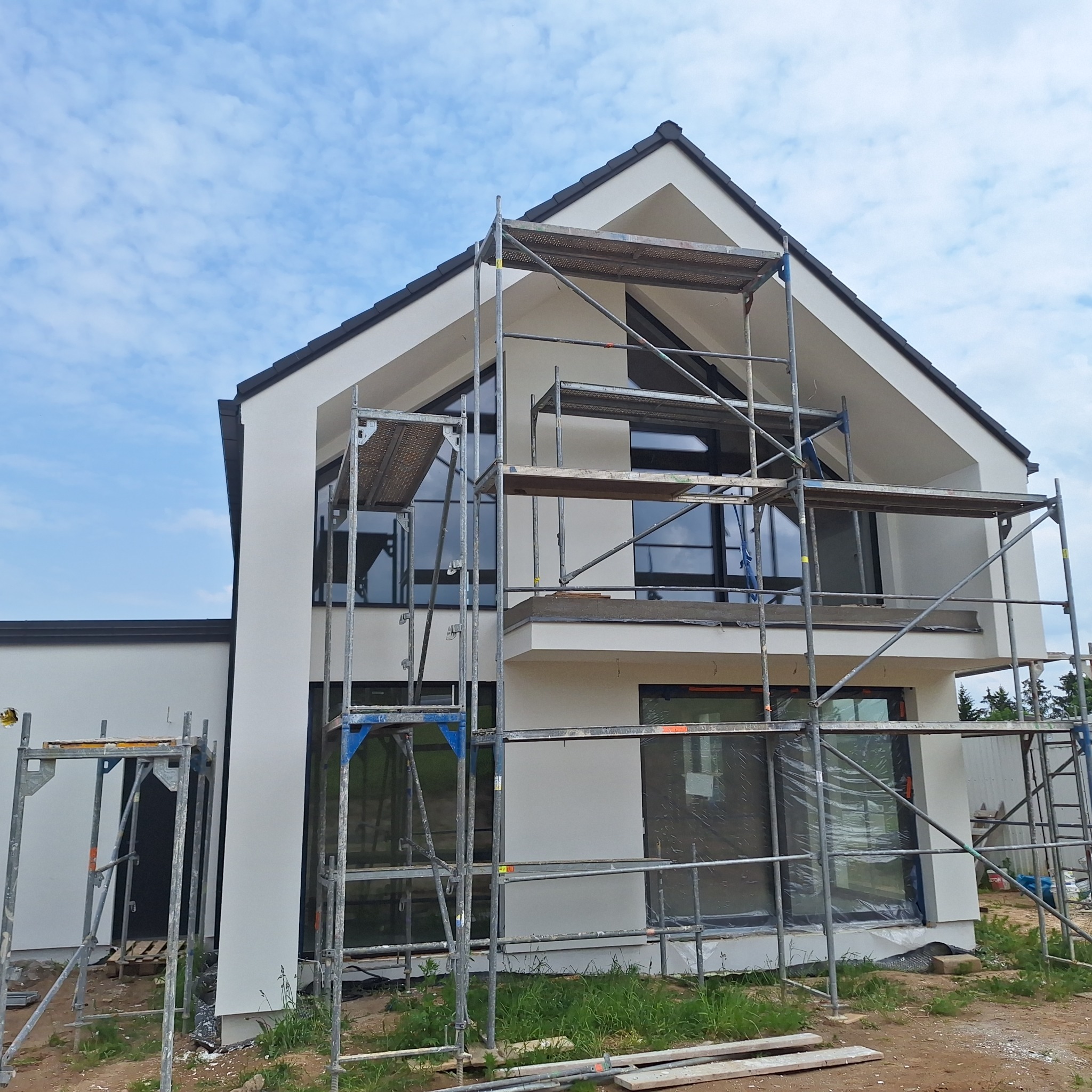 Biała elewacja domu jednorodzinnego w stylu nowoczesnym, z czarną stolarką okienną i dachem, częściowo zasłonięta przez rusztowanie.