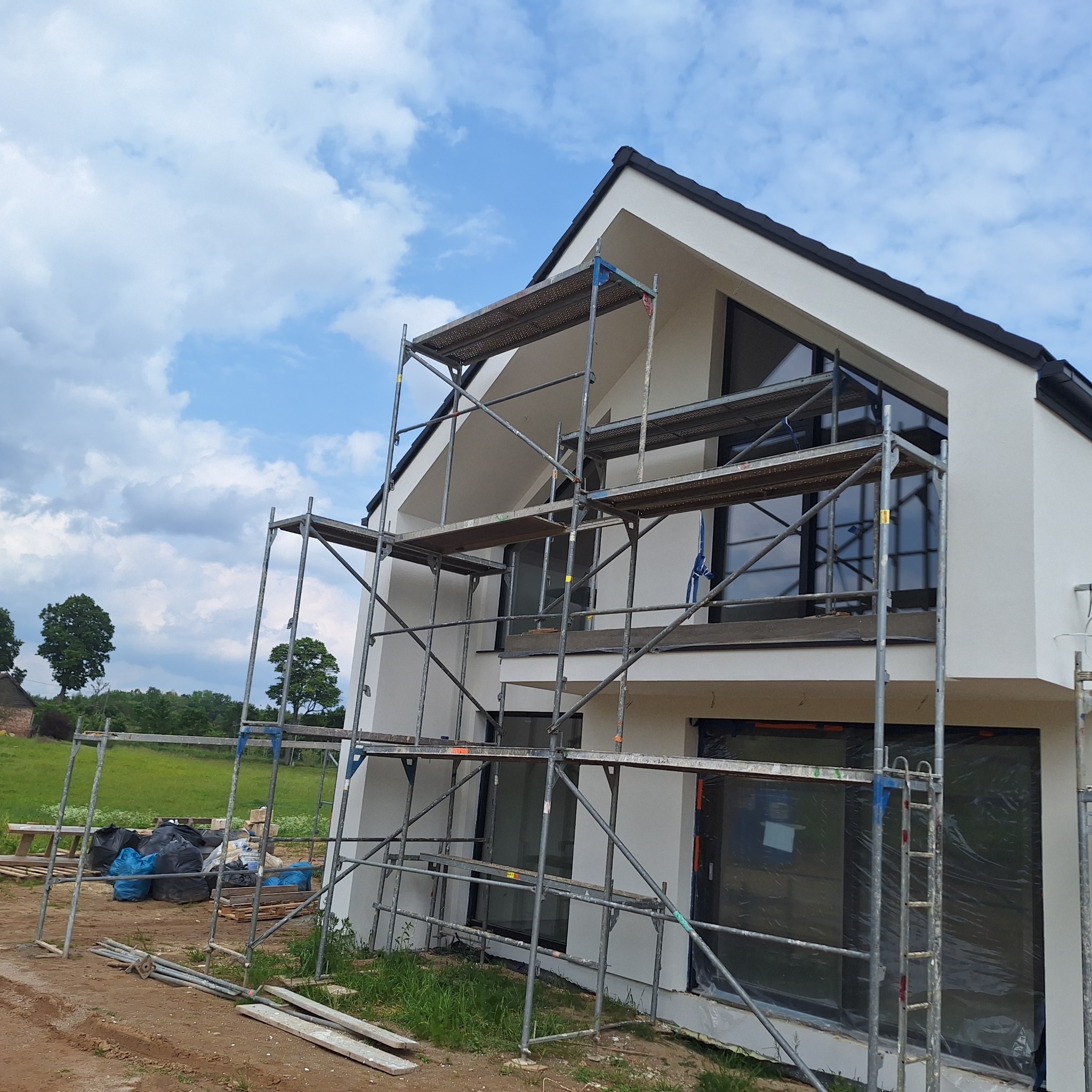 Nowoczesny dom z białą elewacją w trakcie prac. Widoczne rusztowania, okna zabezpieczone folią. W tle zielone pola i błękitne niebo z chmurami.