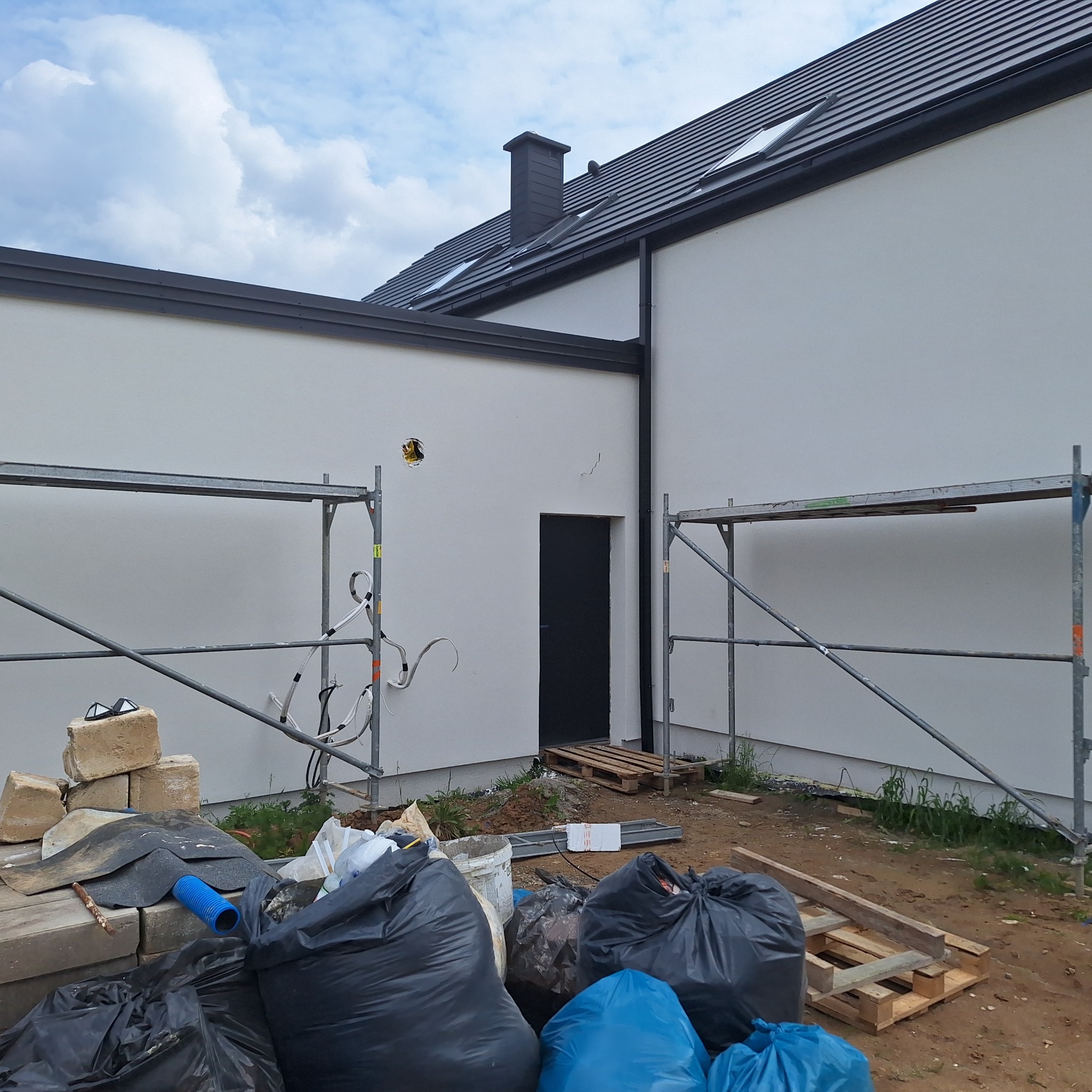 Biała elewacja domu z czarnym dachem i oknami dachowymi, widoczne rusztowania i worki z odpadami budowlanymi. Otwór w ścianie na instalację.