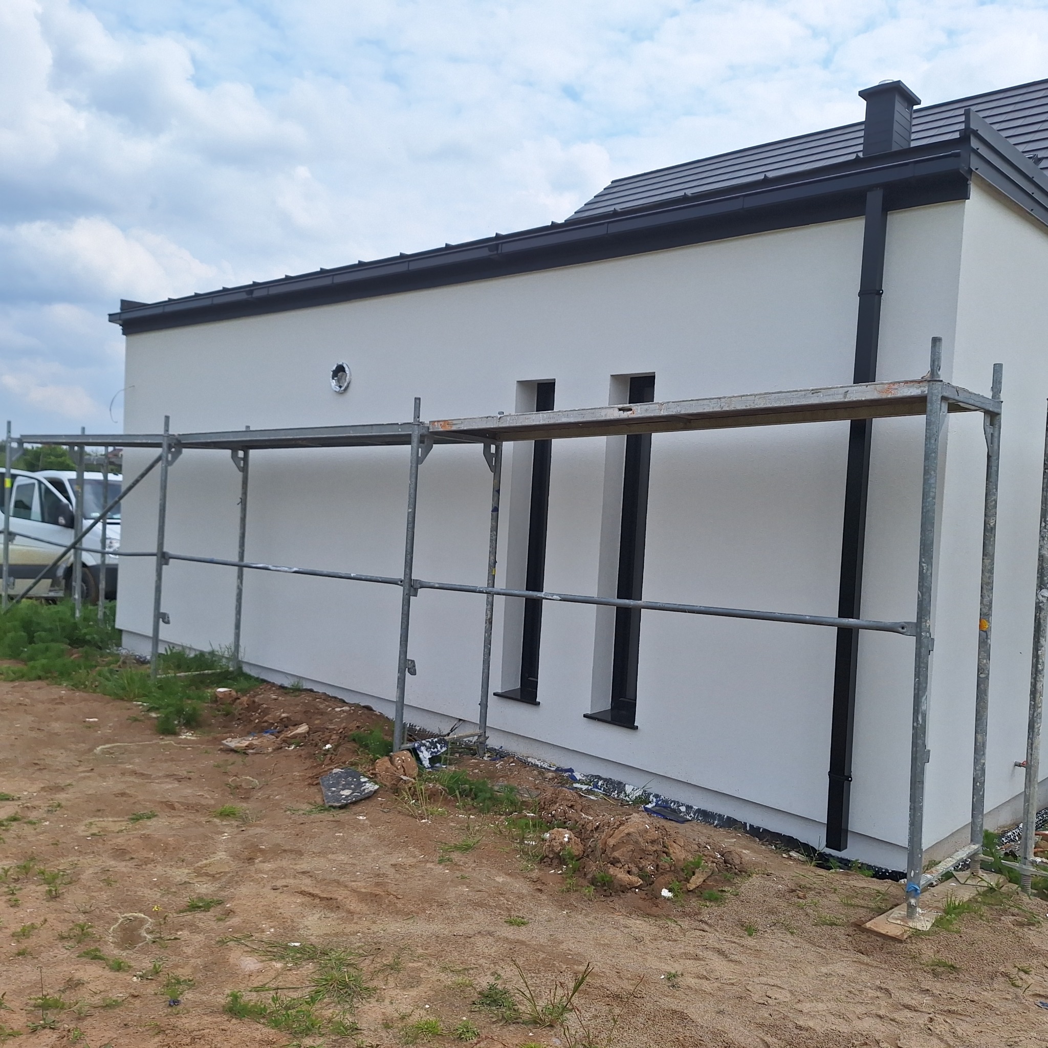 Świeżo otynkowana, biała ściana domu jednorodzinnego z czarnymi rynnami i wąskimi oknami, widoczna z poziomu rusztowania. Częściowo zagospodarowany teren wokół budynku.