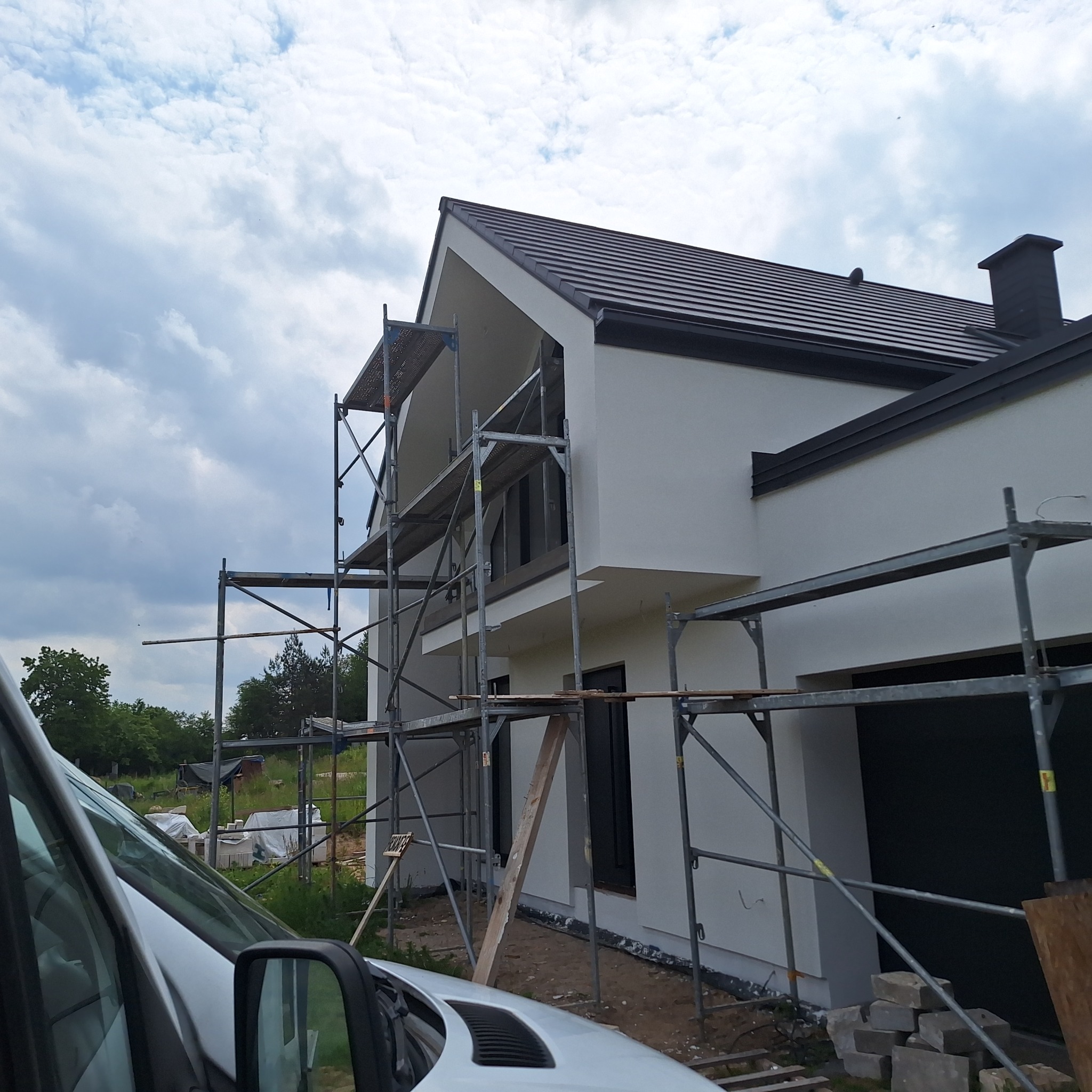 Nowoczesny dom jednorodzinny z białą elewacją w trakcie prac wykończeniowych. Widoczne rusztowania i fragment samochodu dostawczego. Tło: zielone drzewa i pochmurne niebo.