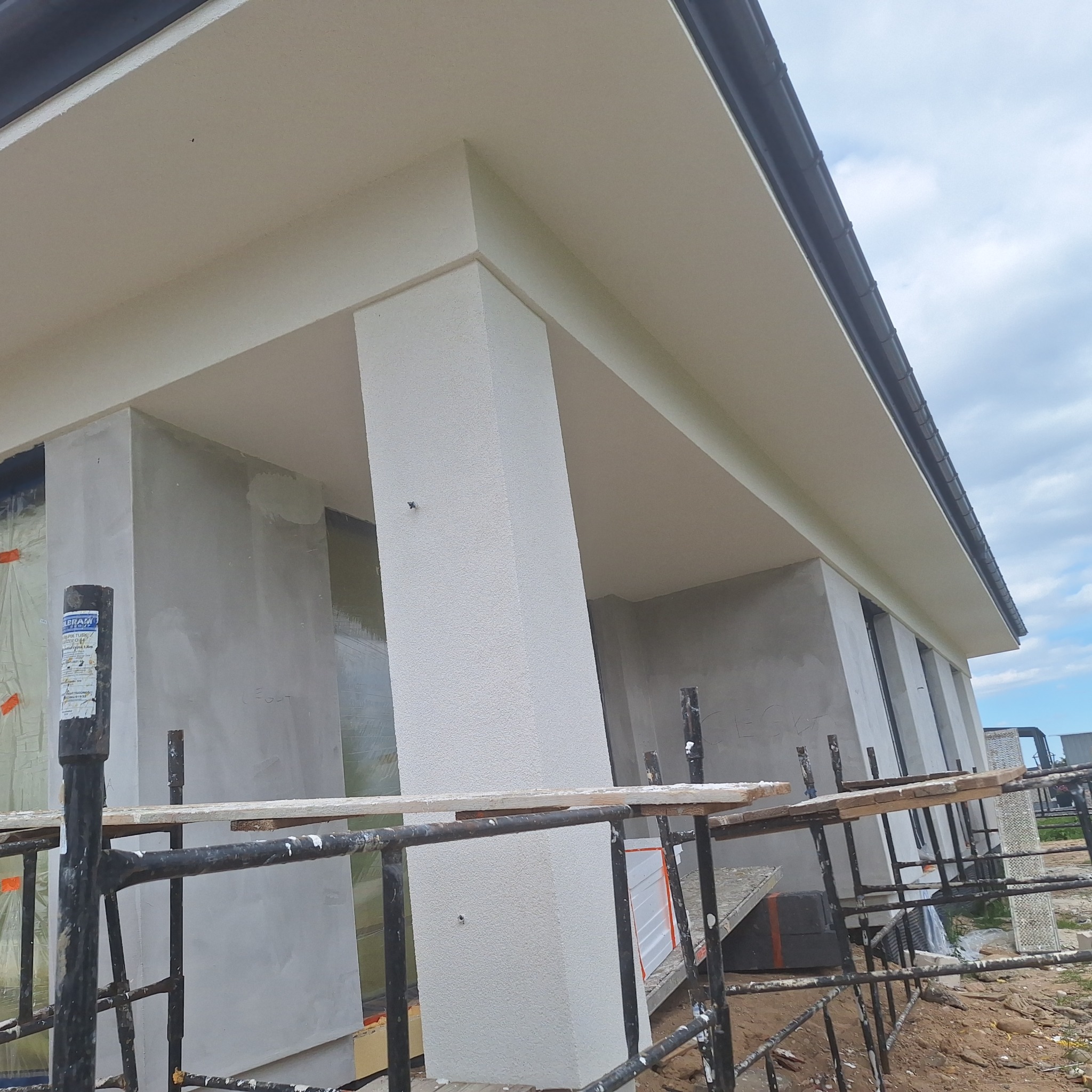 Fragment elewacji domu w trakcie prac wykończeniowych; widoczne rusztowania, otynkowana kolumna i fragment ściany w surowym tynku.