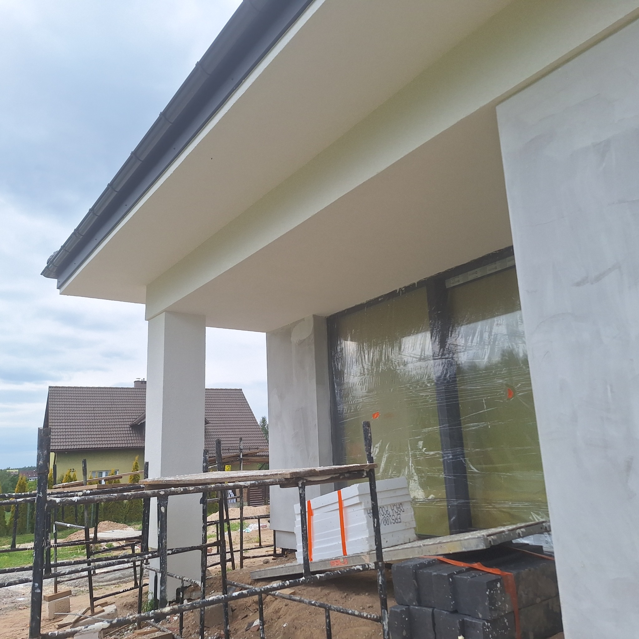 Elewacja domu w trakcie prac: ocieplanie ścian styropianem, zabezpieczone okna folią, widoczne rusztowania i materiały budowlane. Ujęcie z dołu na fragment budynku.