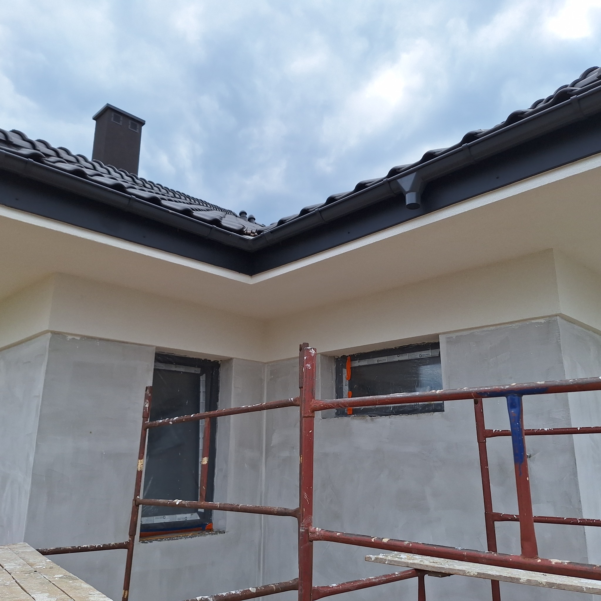 Fragment elewacji domu w trakcie prac wykończeniowych. Widoczne rusztowanie, tynk surowy i pomalowana górna część ściany. Dach z ciemnej dachówki.