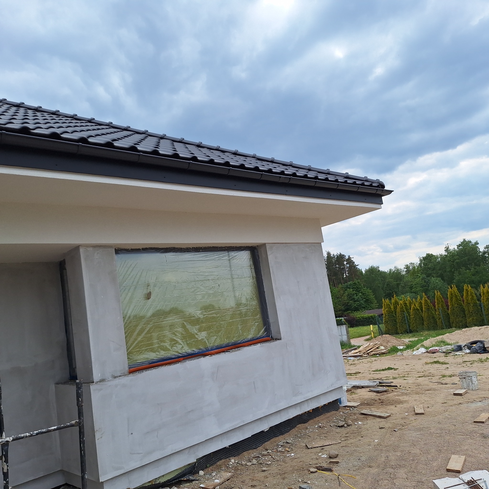Dom jednorodzinny w trakcie tynkowania elewacji. Okno zabezpieczone folią. Widoczne rusztowanie i teren budowy. Dach pokryty czarną dachówką.