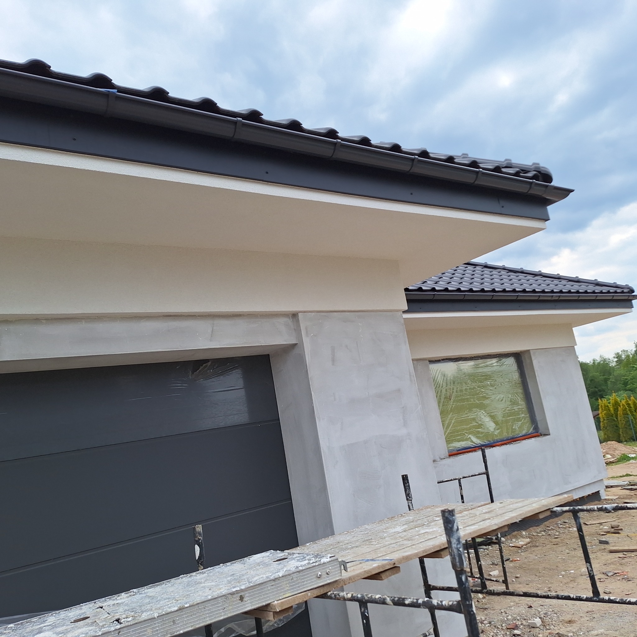 Fragment elewacji domu w trakcie prac wykończeniowych, widoczne rusztowanie, okno zabezpieczone folią. Świeży tynk i pomalowana górna część. Dach z ciemnej dachówki.