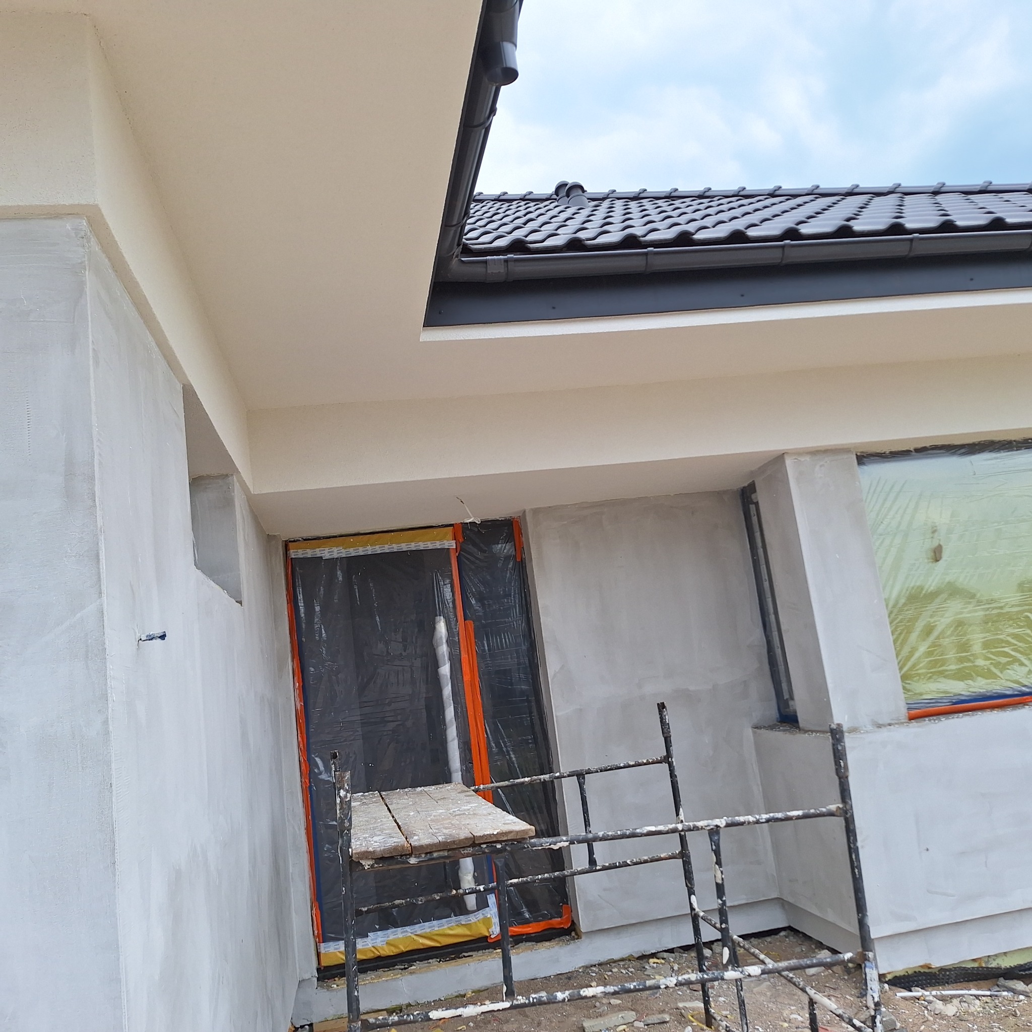 Elewacja domu w trakcie prac wykończeniowych: ściany otynkowane na szaro, okna zabezpieczone folią, widoczne rusztowanie. Dach z ciemnej dachówki.