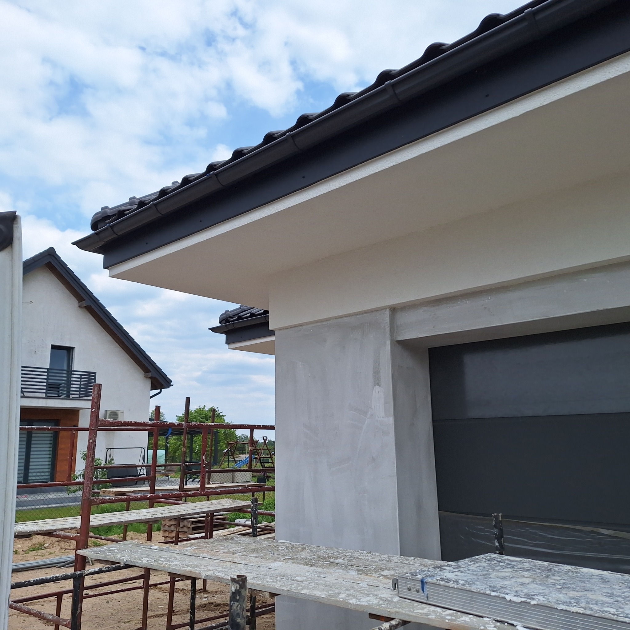 Świeżo otynkowana elewacja domu z ciemną bramą garażową, widoczna z rusztowania. W tle drugi budynek i błękitne niebo z chmurami.