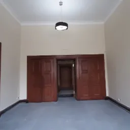 Pusta przestrzeń w odcieniach beżu i szarości z ciemnymi, drewnianymi drzwiami z panelami i listwami przypodłogowymi, oświetlona pojedynczą lampą wiszącą.