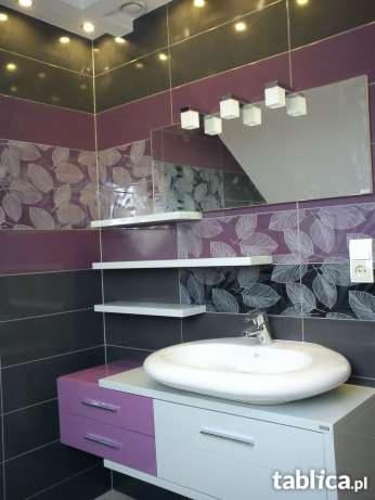 Wykończona łazienka z umywalką w kształcie nerki, lustrem z oświetleniem i półkami na tle płytek w odcieniach fioletu i szarości z motywem liści.