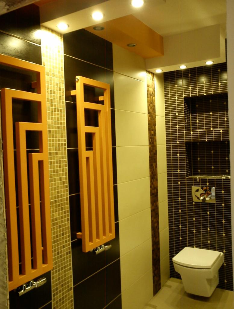 Nowoczesna łazienka z geometrycznymi grzejnikami w kolorze pomarańczowym, mozaiką i ciemnymi płytkami. Podwieszana toaleta i oświetlenie punktowe.