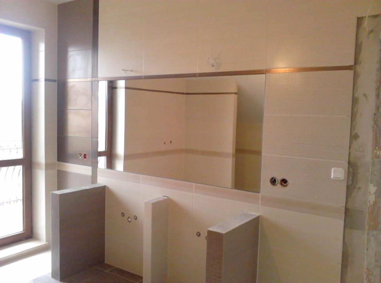 Wykończona łazienka z beżowymi i brązowymi płytkami, lustrem i przygotowanymi otworami na armaturę. Fragment ściany w stanie surowym po prawej stronie.