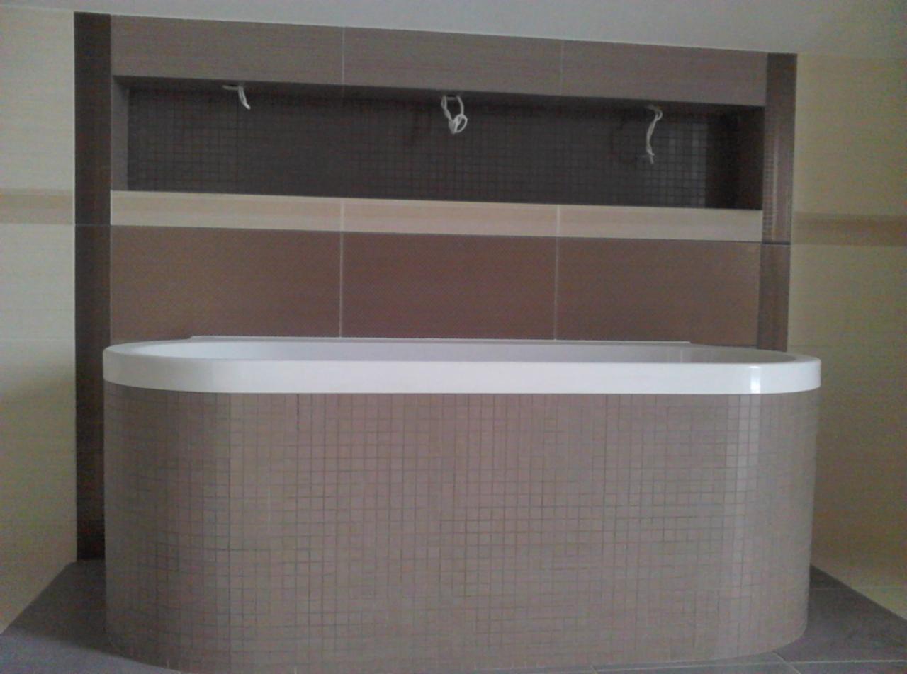 Wykończona łazienka z zabudowaną wanną w mozaikę i półką nad nią, wykończona płytkami w odcieniach brązu i beżu, widok z przodu.